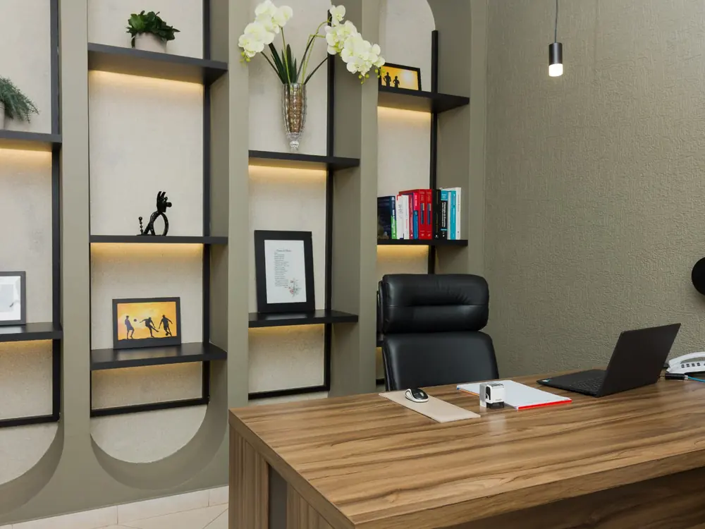 Consultório do Dr. Marcos Sardinha com mesa de atendimento, poltronas para acompanhante e estantes de livros iluminadas